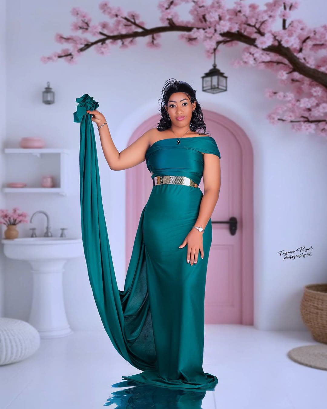Woman in an emerald satin gown holding its train beside a pink arched door and cherry blossom set