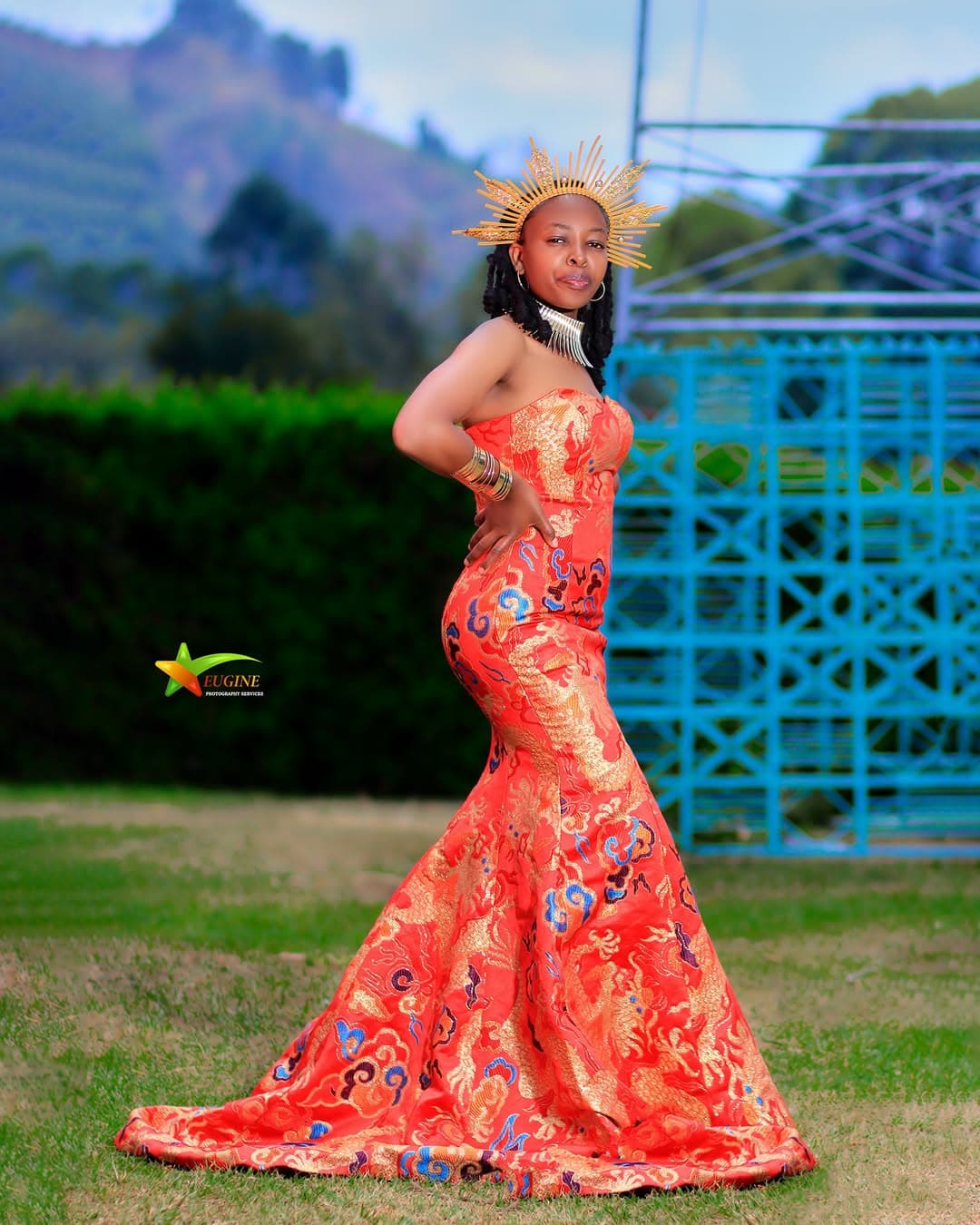 Woman in an embroidered orange mermaid gown and gold crown on a hillside lawn