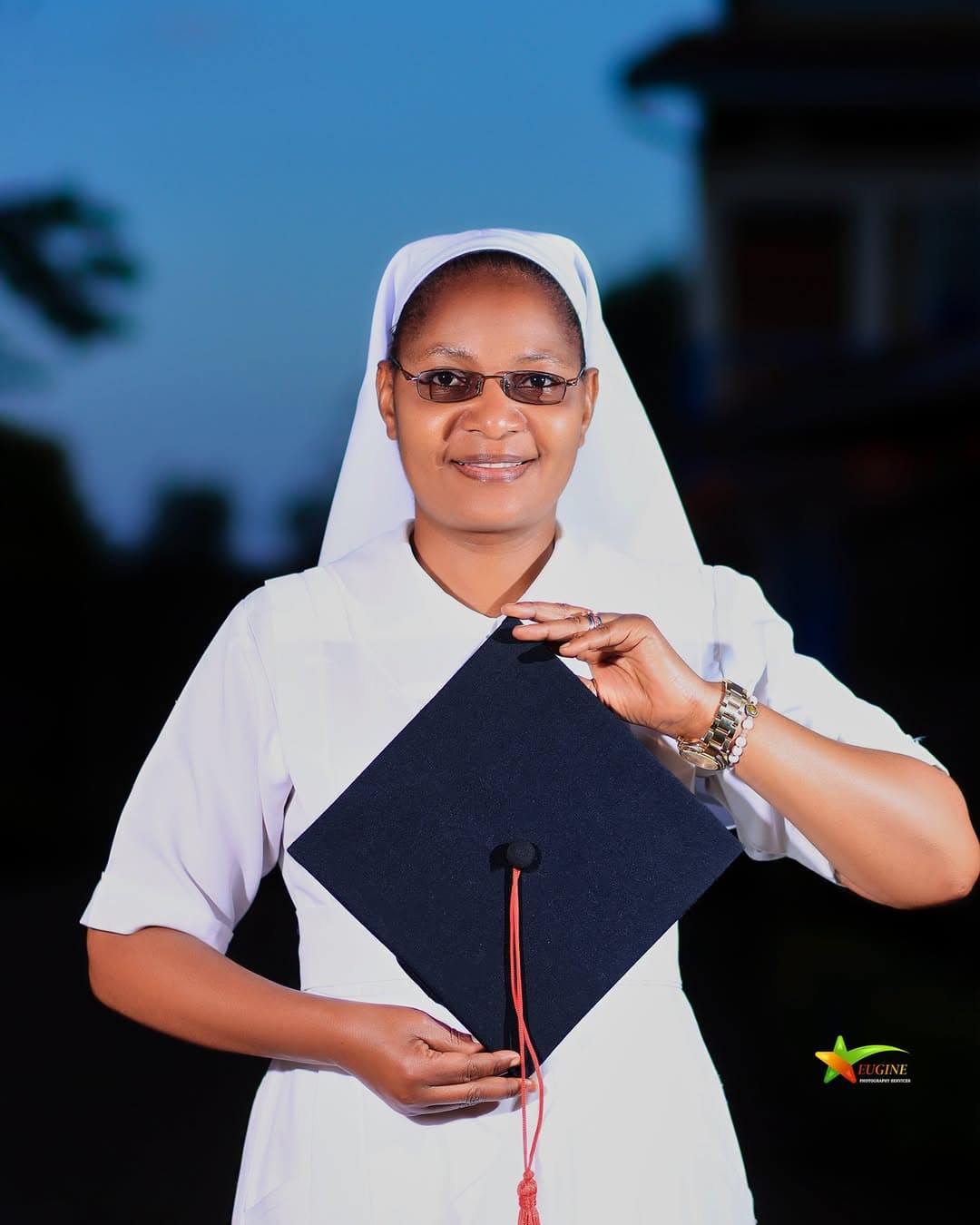 Graduate in a white religious habit holding her mortarboard against a dusk sky