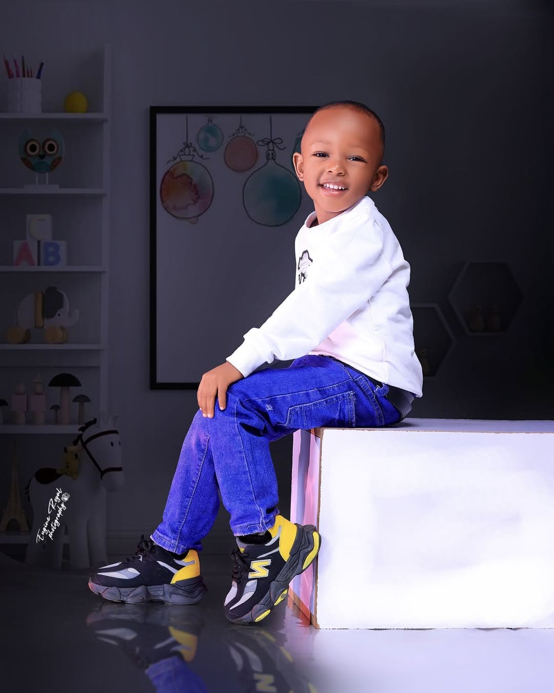 Smiling boy in a white sweatshirt and jeans sitting on a block in a studio set