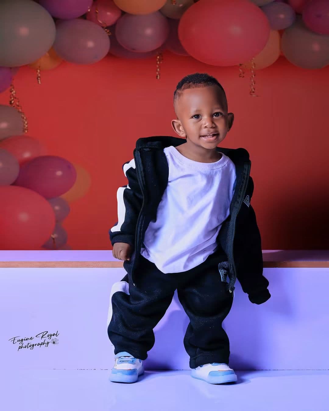 Smiling toddler in a black tracksuit standing in front of pink balloons