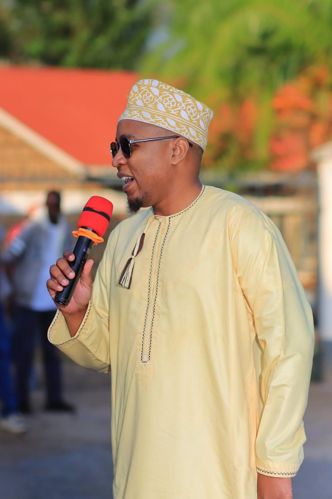 Master of ceremonies in a cream kanzu and kofia speaking into a microphone