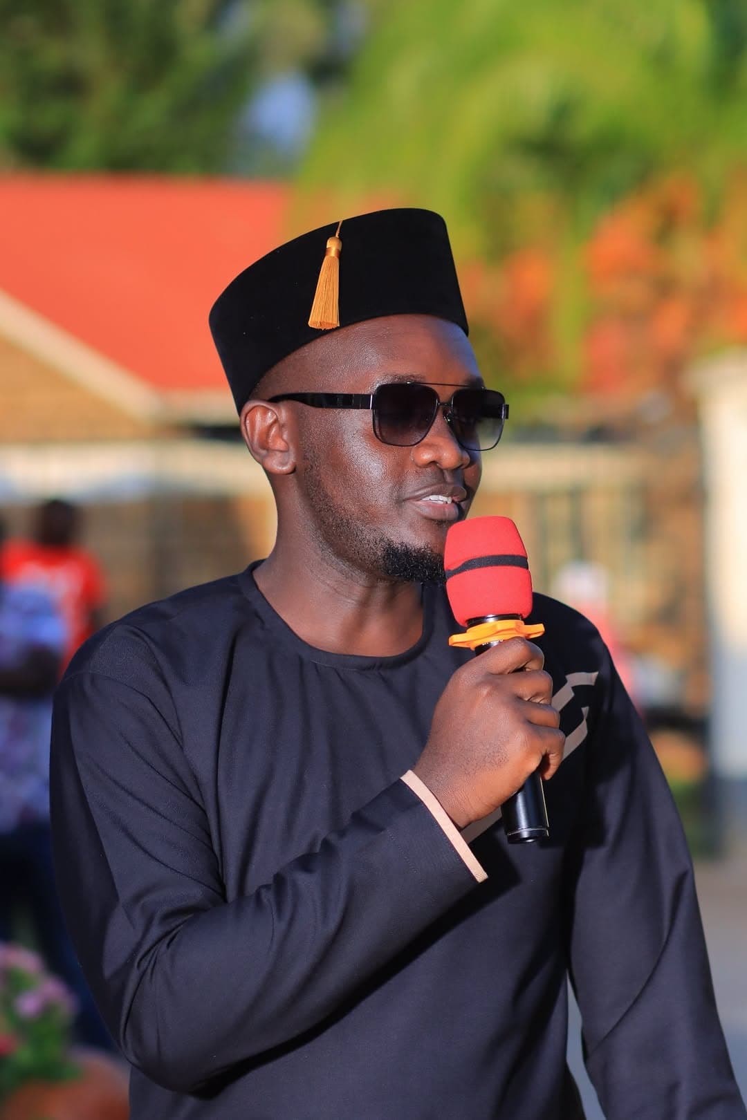 Man in a black outfit, hat and sunglasses speaking into a microphone
