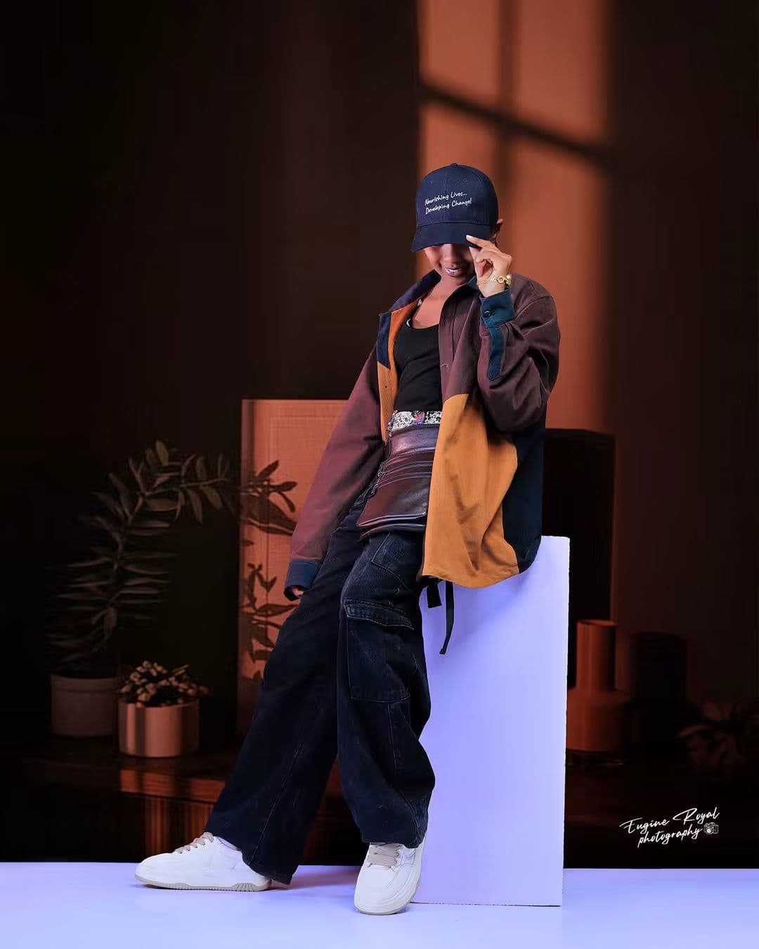 Young man in a cap and layered jacket leaning on a white block in a dim studio set
