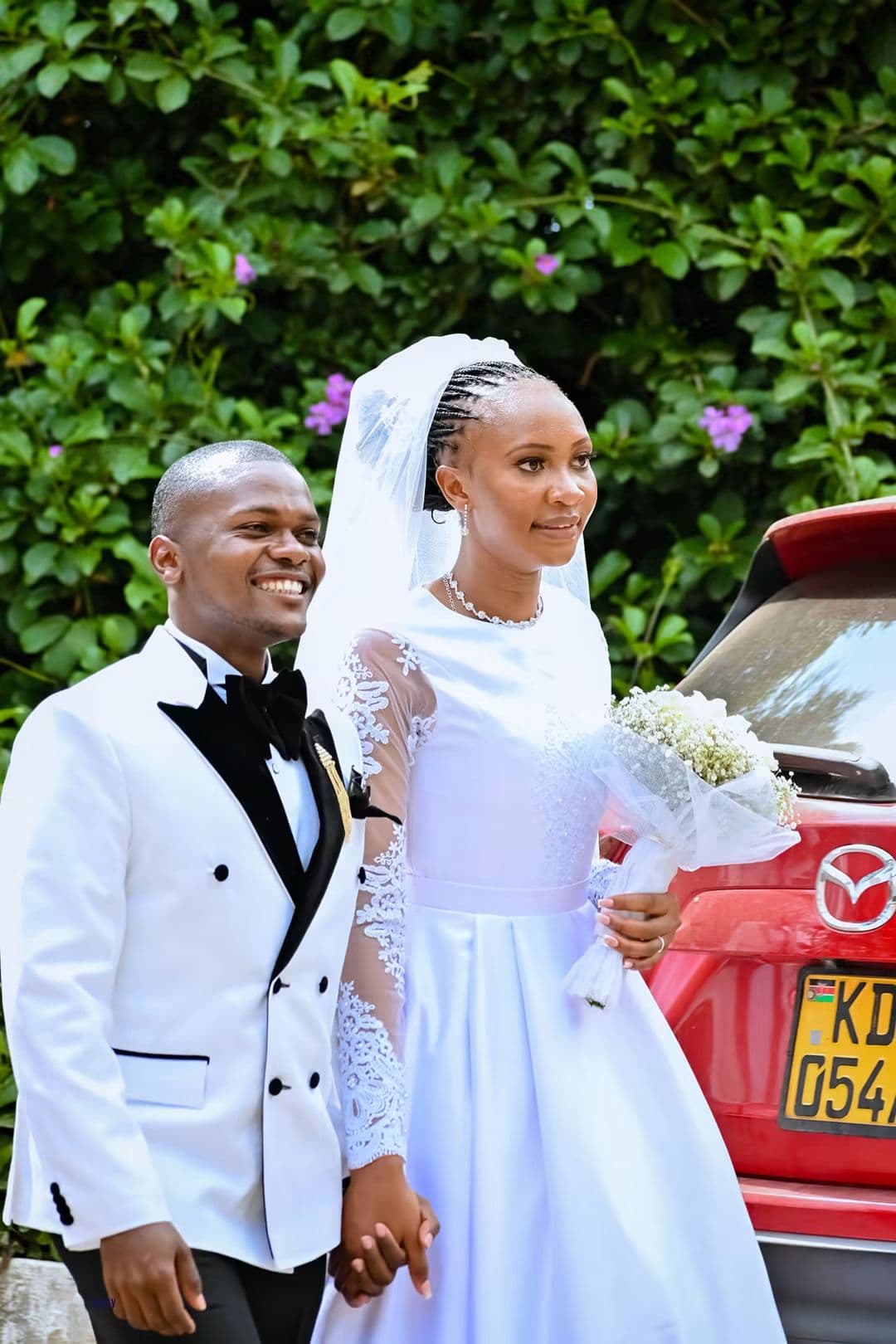 Bride and groom smiling side by side beside a red car