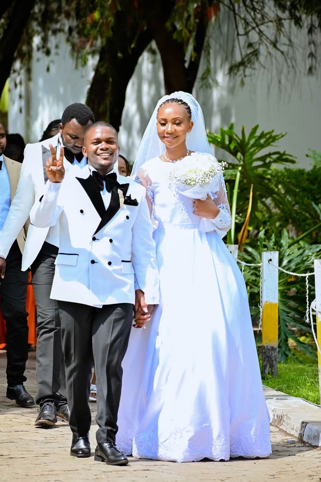 Bride and groom walking along a garden path, the groom raising a peace sign