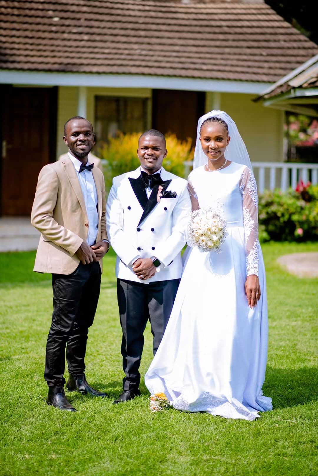 Bride and groom with a groomsman in a beige jacket in front of a cottage