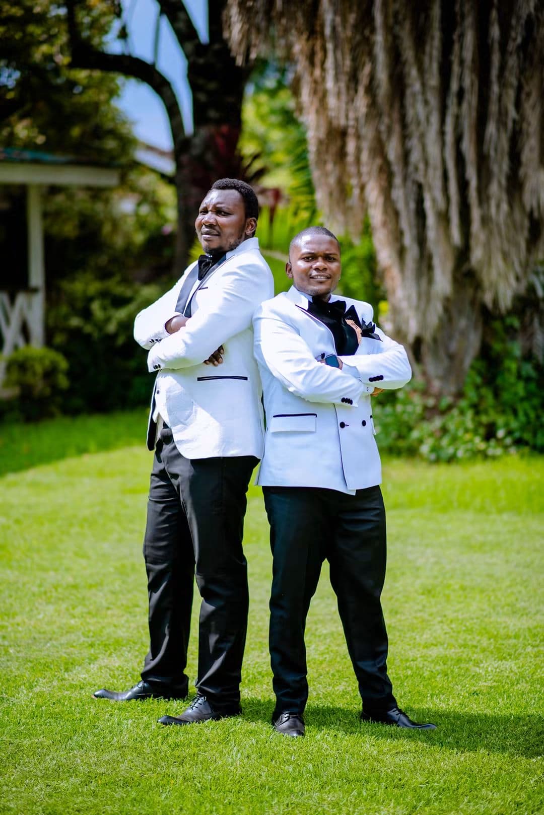 Groom and best man in white tuxedo jackets standing arms crossed under a tree