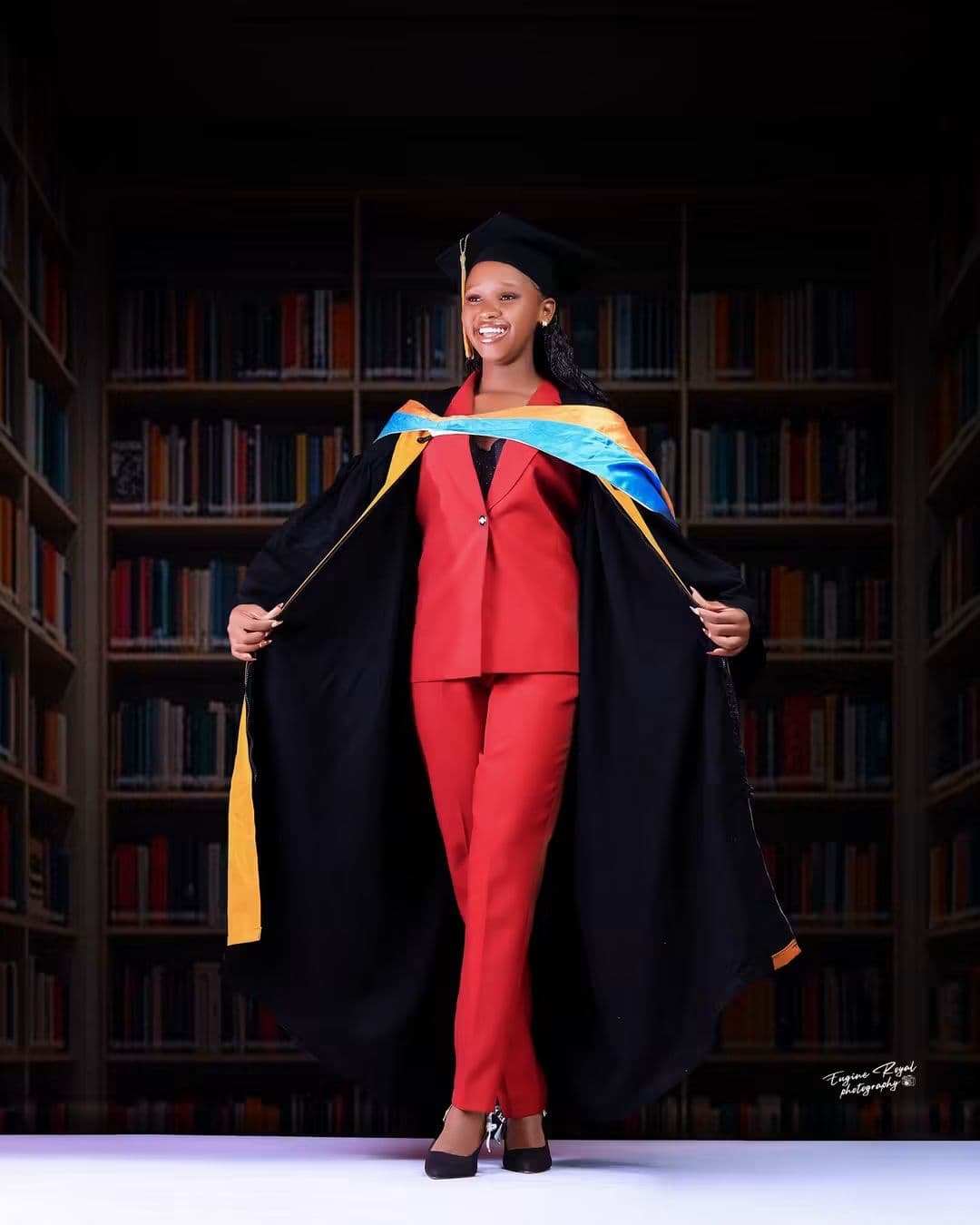 Graduate in a red suit and academic gown standing in a library studio set