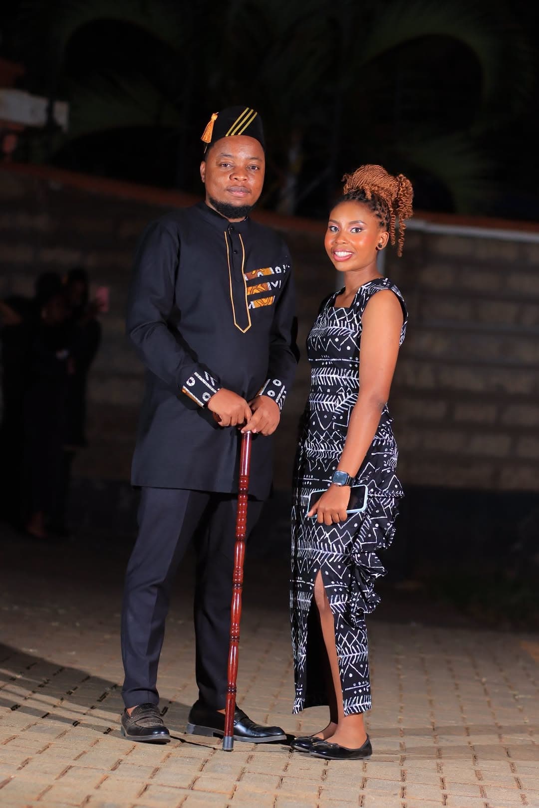 Couple at night, he in black with a walking stick and she in a printed dress