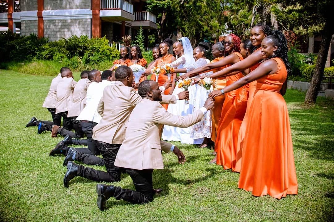 Groomsmen kneeling before the bridesmaids in orange gowns on the lawn